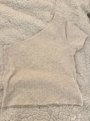 Beige ribbad oneshoulder topp - Säljer en beige ribbad topp med oneshoulder-design. Toppen är croppad och har en kort ärm på ena sidan medan den andra axeln är bar. Perfekt för dig som gillar en stilren och trendig look.