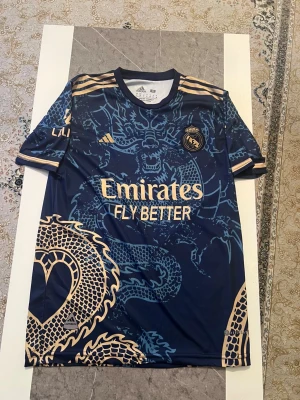 Real Madrid Ronaldo #7 tröja - Snygg Real Madrid fotbollströja med Ronaldo #7 på ryggen, tillverkad av Adidas. Tröjan är mörkblå med guldiga detaljer och har ett unikt drakmönster över hela framsidan och baksidan. Kortärmad med rund hals och tillverkad i lätt polyester för maximal komfort.