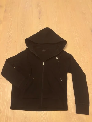 Svart hoodie från Polo Ralph Lauren - Svart hoodie från Polo Ralph Lauren med klassisk vit broderad logga på bröstet. Tröjan har huva med snörning, hel dragkedja framtill och två fickor. Materialet är mjuk bomull och passformen är normal. Perfekt för chill dagar och streetwear.