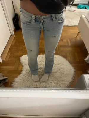 Ljusblå bootcut jeans med slitningar - Snygga ljusblå jeans med bootcut passform och slitna detaljer på framsidan. Jeansen har låg midja, klassiska fem fickor och coola ficklock med knapp bak. Perfekt för dig som gillar en avslappnad och trendig stil.