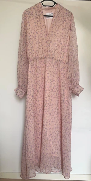 Slected Femme - Långklänning från Selected Femme i ljusrosa med lila blommigt mönster. Klänningen har v-ringning, långa ärmar med knapp vid manschetten och ett lätt, luftigt tyg. Obs: Alla plagg säljs endast fram till 28/12 