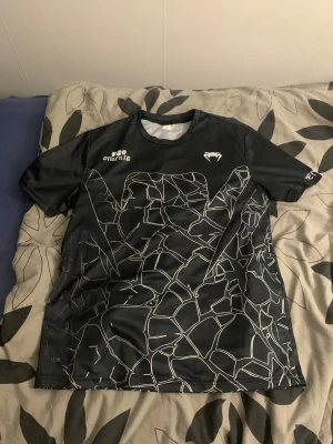 Svart tröja  Omerta S - Cool svart tröja från  m Omerta-tryck och unikt vitt grafiskt mönster som liknar spruckna linjer över hela tröjan. Kortärmad modell i lätt och ventilerande polyester, perfekt för träning eller match. Snyggt tryck både fram och bak.