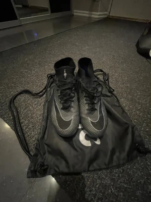 Nike Zoom Mercurial fotbollsskor - Säljer ett par svarta Nike Zoom Mercurial Elite fotbollsskor med högt skaft och snörning. Skorna har en rund tå och platt sula med dobbar för konstgräs. Materialet är syntet med en cool textur och silverfärgade detaljer. Medföljer original Nike-påse.
