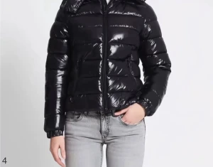 Svart pufferjacka med huva - Säljer en svart, glansig pufferjacka med huva och dragkedja framtill. Jackan har en quiltad look med breda kanaler och långärmade armar. Perfekt för kalla dagar när du vill ha en stilren och chill vibe. Passar dig som gillar en klassisk streetstil.