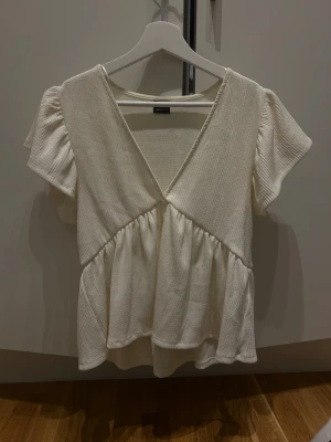 Vit omlott blus från Gina Tricot - Säljer en söt vit blus från Gina Tricot med omlott-design och volangdetaljer. Blusen har korta, lätt puffade ärmar och djup v-ringning. Materialet är mjukt och har en lätt struktur som ger en skön känsla. Perfekt för dig som gillar romantisk och feminin stil.