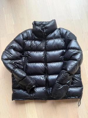 Svart Moncler pufferjacka dam - Säljer en svart pufferjacka från Moncler med hög krage och diskret logotyp på ärmen. Jackan har dragkedja framtill, två sidofickor med dragkedja och justerbar dragsko nedtill. Materialet är glansigt och dunfyllt, perfekt för kalla dagar.