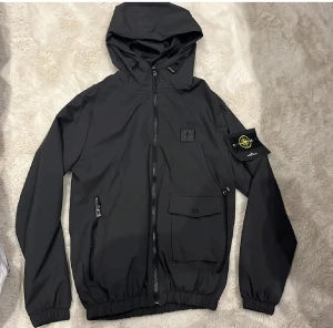 Svart vindjacka Stone Island S - Svart vindjacka från Stone Island med huva, dragkedja framtill och två fickor med zip och knapp. Klassisk patch med logga på vänster ärm och diskret logomärke på bröstet. Jackan har elastiska muddar och är tillverkad i lätt syntetmaterial.