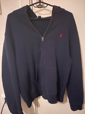 Marinblå hoodie från Polo Ralph Lauren  - Snygg marinblå hoodie från Polo Ralph Lauren i storlek XL. Tröjan har huva, dragkedja framtill och en röd broderad logga på bröstet. Har knappt använt den, riktiga kostar 1225 säljer för mycket mindre 