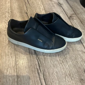 Marin blå sneakers från Axel Arigato - Stilrena svarta sneakers från Axel Arigato med vit sula och diskret guldfärgad logga på sidan. Skorna har en minimalistisk design utan snörning och bred elastisk panel över vristen. Perfekta för en clean och modern look. Knappt använda. Pris kan diskuteras