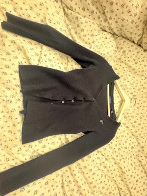 Mörkblå kofta från GANT i ull XS - Mörkblå kofta från GANT i mjuk ull, med v-ringad hals och knappar framtill. Liten broderad GANT-logga med sköld på bröstet. Långärmad och klassisk passform, perfekt för lager-på-lager under kyliga dagar. Har krymt i tvätten så kommer tyvärr vara mindre än vad den ska vara så jag föreslår att man går ner en storlek, alltså XXS.