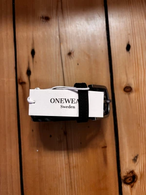 blå och grått flätat bälte OneWear - Säljer ett snyggt flätat bälte från OneWear Sweden i blå och grått med inslag av vitt. Bältet har ett stilrent metallspänne och detaljer i svart skinn vid ändarna. Perfekt för att ge outfiten en cool touch.