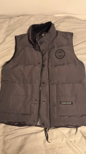 Mörkgrå dunväst med svart Canada Goose logga - Mörkgrå canada goose dunväst med svart label. En väldigt snygg Dunväst som värmer riktigt bra. Jättebra skick, kontakta ifall du är intresserad.
