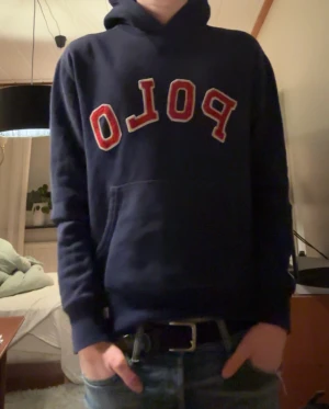 Mörkblå hoodie från Polo Ralph Lauren - Säljer en mörkblå hoodie från Polo Ralph Lauren med stor röd och vit POLO-text på bröstet. Tröjan har huva, känguruficka och ribbade muddar. Perfekt för dig som gillar stilrena kläder! 
