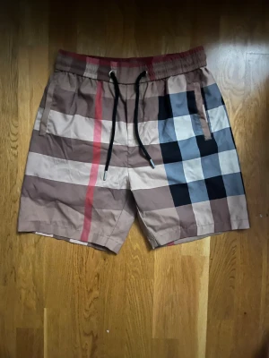 Rutig Burberry shorts med snörning - Säljer ett par snygga Burberry shorts med klassiskt rutigt mönster i beige, svart, vit och röd. De har elastisk midja med svart snörning och sidofickor. Perfekta för sommaren och har en chill loose fit. Materialet känns som bomull eller polyester.