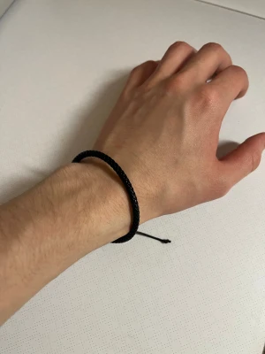 Flätat armband  - Stilrent svart armband med flätad design och justerbar knytning. Perfekt för dig som gillar minimalistiska accessoarer och vill ha något enkelt men snyggt runt handleden. Passar både tjejer och killar och är lätt att matcha med andra smycken.