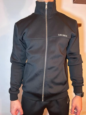 Svart track sett från Les Deux - Svart track sett från Les Deux med hög krage och hel dragkedja framtill. Jackan har ribbade muddar, två fickor och diskret logga på bröstet. Snygg och stilren modell i mjukt syntetmaterial, perfekt för en sportig look. Både tröjan och byxorna är storlek S