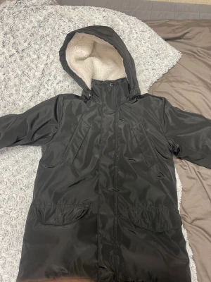 Svart parkajacka från On The Peak - Svart parkajacka från On The Peak med huva och mysigt vitt teddyfoder på insidan. Jackan har flera fickor framtill, dragkedja och tryckknappar. Perfekt för kalla dagar och har en sportig vibe.