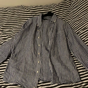 Randig blå skjorta från Massimo Dutti - Snygg skjorta från Massimo Dutti med blå och vita vertikala ränder, klassisk krage och knappar framtill. Skjortan har lång ärm med mörkblå armbågslappar och en bröstficka. Materialet är lätt och känns som bomull, perfekt för en chill och stilren look.