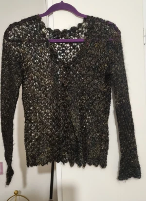Svart glittrig kofta med knappar. Handstickat av ull. Kan passa som S/M. - Säljer en svart glittrig kofta med lång ärm och knappar framtill. Koftan är stickad i ett genombrutet mönster och har en lätt glansig finish som ger en cool look. Perfekt att slänga över en topp för extra stilpoäng.