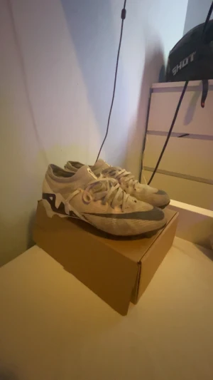 Nike mecurial elite fotbollsskor  - Säljer ett par Nike fotbollsskor i beige med svarta detaljer och logga. Skorna har snörning och en låg profil, med en platt och mönstrad sula för bra grepp på planen. Ovandelen är i syntetmaterial och mesh för lätt känsla och ventilation. Skorna är i storlek 41 men Nike fotbollsskor brukar vara små i storleken så känns som 40. Det finns lite slitage på skon som syns på bild 5 men det lilla hålet påverkar inte skornas effekt.
