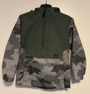 Vindjacka strl 110 - Snygg vindjacka från Urban Classics i grön och grå camouflage med halv dragkedja framtill och huva. Jackan har elastiska muddar vid ärmslut och en cool patch på framsidan. Köpt på Lager157