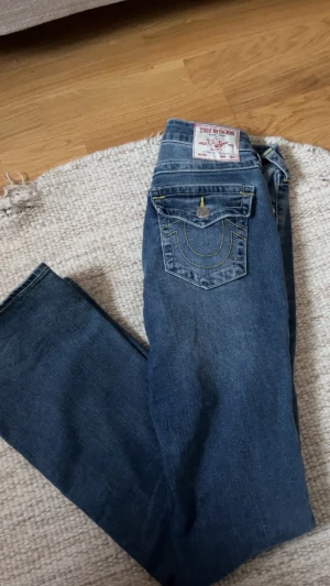 True Religion Becca Bootcut Jeans - Säljer ett par snygga True Religion Becca jeans i klassisk blå tvätt med bootcut. De har gul kontrastsöm, silverfärgade knappar och ficklock bak med den ikoniska loggan. Midwaist passform och tillverkade i mjukt jeanstyg för en chill look.