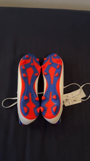 Adidas Copa Pure 2 Club Fx fotbollsskor - Säljer ett par Adidas Copa Pure 2 Club Fx fotbollsskor i vitt syntetmaterial med blåa detaljer och röda Adidas-loggor. Skorna har snörning, rund tå och en yttersula med röda och blå dobbar för gräsplan. Klassisk design med tre blå ränder på sidan.