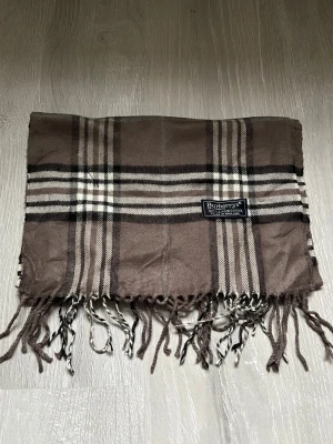 Brun rutig halsduk från Burberrys - Snygg halsduk från Burberrys i klassiskt rutigt mönster med bruna, beige och svarta toner. Gjord i 100% lammull och har fransar i ändarna som ger en chill vibe. Perfekt att slänga över axlarna när det är kallt ute.