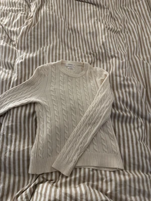 Vit kabelstickad tröja från Reserved - Snygg vit kabelstickad tröja från Reserved med rund hals och långa ärmar. Tröjan har ett klassiskt flätmönster och ribbade muddar vid ärmslut och nederkant. Storlek M