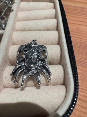 Silverfärgad ring med jokerdesign - Unik och chunky ring i silverfärgad metall med ett detaljerat motiv av en joker eller narr. Ringen har en cool och edgy vibe med ansiktet och de typiska narrspetsarna som sticker ut. Perfekt för dig som gillar statement-smycken och vill sticka ut.