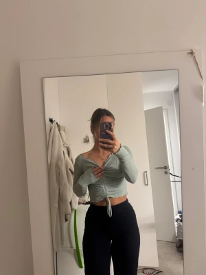 Ljusgrön croppad omlottopp - Säljer en ljusgrön croppad omlottopp med långa ärmar och snygg snörning framtill. Toppen har ribbad struktur och v-ringad hals, vilket ger en fräsch och trendig look. Perfekt att styla med höga jeans eller kjol.