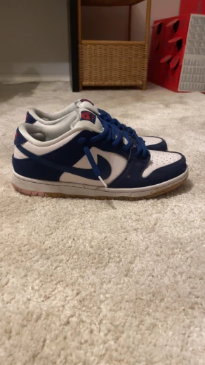 Nike SB Dunk Low blå och vita sneakers - Nike SB Dunk Low sneakers i blått och vitt med klassisk låg siluett. Skorna har blå snörning, perforerad tåbox och gummisula i brunt. Märket syns tydligt på plösen med röd logga. Ovandelen är i syntet och skinn för en snygg streetstil.