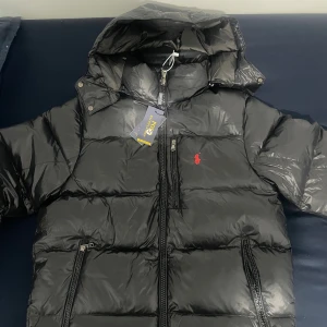 Svart pufferjacka från Polo Ralph Lauren - Svart pufferjacka från Polo Ralph Lauren med röd broderad logga på bröstet. Jackan har huva, flera dragkedjefickor och RL-detaljer på dragkedjan. Materialet är glansigt syntet och jackan är quiltad för extra värme. Perfekt för kalla dagar.