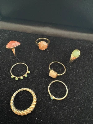 Set med sex guldiga ringar med stenar - Säljer ett set med sex olika guldiga ringar. Några har orange och gröna stenar i olika former, en är tvinnad och chunky, andra har små detaljer som geometriska former och pärlliknande stenar. Perfekt för dig som gillar att mixa och matcha smycken med färg och cool design.
