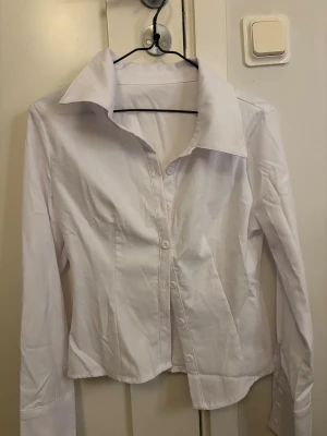 Vit klassisk figur sydd skjorta med knappar - Snygg vit skjorta med klassisk krage och knappar framtill. Skjortan har lång ärm med bred manschett och är tillverkad i ett slätt bomullstyg. Passformen är normal och designen är stilren, perfekt för dig som gillar en clean look.