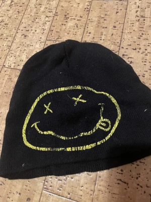 Svart Nirvana mössa med gul logga - Svart stickad mössa med Nirvanas ikoniska smiley-ansikte i gult på ena sidan och broderad gul Nirvana-text på andra sidan. Perfekt för dig som vill visa din kärlek till grunge och bandet Nirvana. Mjuk och skön passform som håller dig varm.