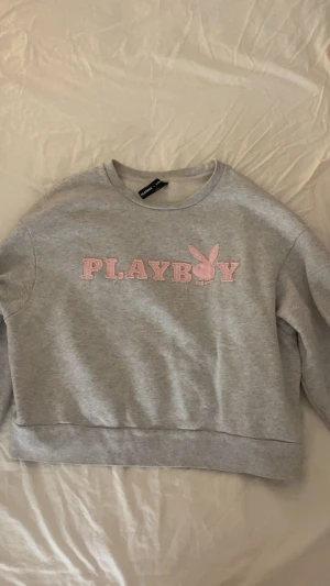 Grå sweatshirt från Playboy med rosa tryck - Säljer en grå sweatshirt från Playboy med rosa broderat PLAYBOY-tryck och kaninlogga på bröstet. Tröjan har rund hals, långa ärmar och ribbade muddar vid ärmslut och nederkant. 