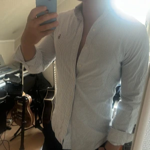 Randig skjorta från Polo Ralph Lauren - Säljer en klassisk skjorta från Polo Ralph Lauren med blå och vita smala ränder. Skjortan har lång ärm, knappar framtill och den ikoniska broderade loggan på bröstet. Tillverkad i mjuk bomull och har en snygg, normal passform.