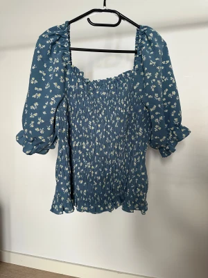 Topp Vila  - Blommig topp i blått med små vita och gula blommor. Toppen har puffiga korta ärmar med volangkant och smockad kropp.   Obs: Alla plagg säljs endast fram till 28/12 
