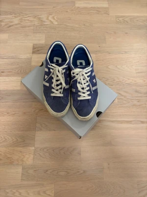 Converse CONS sneakers i mocka - Säljer ett par blå Converse CONS sneakers med vit sula och vita snören. Skorna har klassisk stjärndesign på sidan och är tillverkade i mocka för en cool och retro vibe. Perfekta för dig som gillar streetstyle och vill ha något som sticker ut. Skorna är bra överlag utöver yt smutset men det går bort enkelt med lite skrubbande.