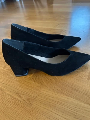 Svarta mockapumps från Tamaris - Snygga svarta pumps från Tamaris i mjuk mocka med spetsig tå och låg klack. Skorna har en klassisk och stilren form som ger en elegant vibe. Perfekta för dig som gillar minimalistisk design och vill ha ett par bekväma klackar med platt sula.
