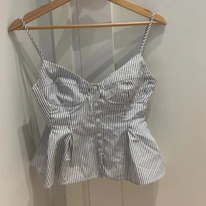 Randig korsett-topp från Zara - Säljer en eftertraktad snygg vit och svart randig korsett-topp från Zara i storlek S. Toppen har smala axelband, knappar framtill och en figurnära passform med utsvängd nederdel. Perfekt för dig som gillar trendiga detaljer och en feminin siluett.
