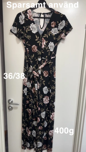 Svart blommig jumpsuit med knyt - Svart jumpsuit från Happy Holly med v-ringning och blommigt mönster i beige, vitt och rosa. Jumpsuiten har korta ärmar och ett snyggt knytband i midjan som ger en fin siluett. Perfekt för dig som gillar romantiska detaljer och mjuka material.🌸 Sparsamt använd. Storlek 36/38