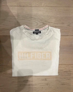 Vit Hilfiger t-shirt med tryck - Vit t-shirt från Tommy Hilfiger med klassiskt tryck i ljusbeige på bröstet. Rund halsringning med diskret röd och blå detalj på insidan av kragen. Kortärmad och tillverkad i mjuk bomull, perfekt för en clean och enkel stil.