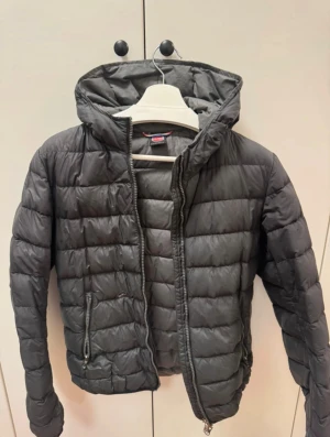 Svart Colmar pufferjacka med huva - Svart pufferjacka från Colmar med huva och två sidofickor med dragkedja. Jackan har ett quiltat mönster och Colmar-logga på ärmen. Perfekt för kalla dagar och har en snygg, sportig vibe. Stängs med dragkedja framtill.väl använd 