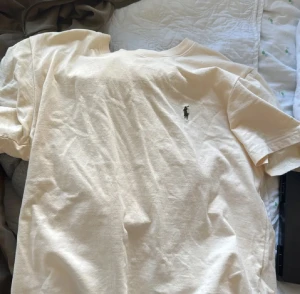 Beige t-shirt från Polo Ralph Lauren - Snygg beige t-shirt från Polo Ralph Lauren med klassisk rund hals och korta ärmar. T-shirten har den ikoniska Polo-loggan broderad på bröstet och är gjord i mjuk bomull som känns skön mot huden. Perfekt för en clean och stilren look. Aldrig använd nästan ny.