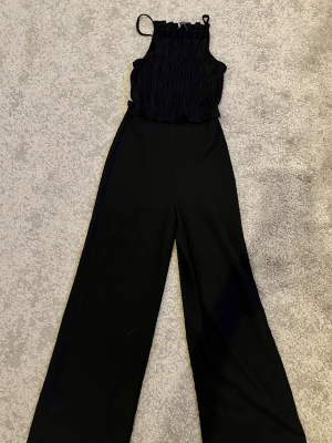 Svart byxdress från Bershka s/xs - Svart byxdress från Bershka med smockad topp och tunna axelband. Byxorna har en loose och wide passform med hög midja och raka ben. Materialet är mjukt och lätt, perfekt för dig som gillar stilrena och bekväma plagg.