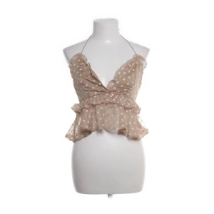 Beige omlott topp med volang från EillyBazar - Supersöt beige omlott topp från EillyBazar i storlek S. Toppen har vita prickar, volangkant och är ärmlös med smala halterneck-band. Materialet är lätt och transparent polyester, perfekt för varma dagar. V-ringad och croppad modell som ger en trendig vibe.