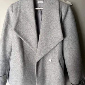 Grå kappa med bälte från Lascana - Snygg grå kappa från Lascana med bred krage och bälte i midjan. Jackan har en stilren design, raka ärmar och är tillverkad i ett mjukt ull-liknande material. Perfekt för dig som vill ha en clean och modern look. Passar xs/s