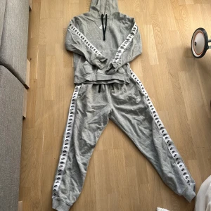 Grå Nike hoodie med logoband - Säljer en grå Nike hoodie med svarta dragsnören och vita band med svarta Nike-loggor längs ärmarna. Tröjan har huva och känguruficka, tillverkad i mjuk bomullsblandning. Perfekt för chill eller träning.